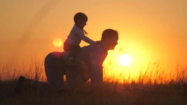 Pappa leker med sin dotter i parken på gräs i solnedgångens strålar. Far rider barnet på ryggen, han sitter bredvid far, jublar. Pappa och barnet leker utomhus. Lycklig familj — Stockvideo