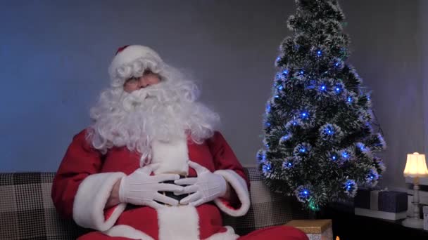 Santa Claus is sitting on couch next to Christmas tree and laughing joyfully. Santa Claus gives audience a gift of fun, laughter, joy. Merry Christmas. Family holiday and celebration. — Stock Video