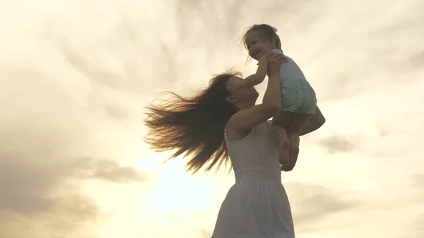 Mutlu anne ve küçük sağlıklı çocuk parkta güneş ışığında doğada sarılıyorlar. Annem sevgili sağlıklı kızını kollarıyla gökyüzüne fırlatıyor. Aile tatilde. Anne ve bebek gün batımında.