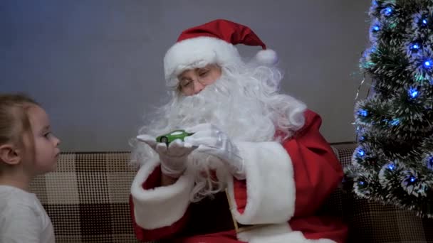 Papai Noel se senta no sofá com uma menina, Papai Noel deu um presente para carro de brinquedo, criança se alegra e abraça o avô mágico. férias e celebração. Família crianças férias de inverno. Feliz noite de Natal. — Vídeo de Stock