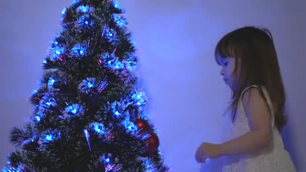 Criança decora a árvore de Natal com bolas de Natal. Criança pequena joga pela árvore de Natal no quarto das crianças. Filha examina guirlanda na árvore de Natal. Infância feliz, férias em família — Vídeo de Stock