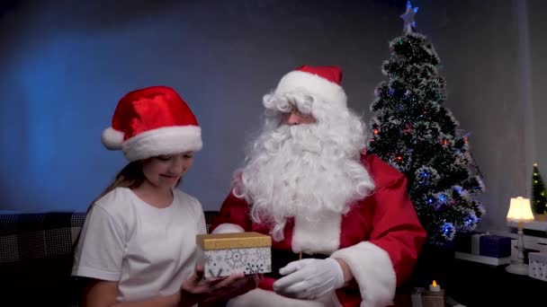 Feliz Nochebuena. Santa Claus da un regalo a una niña, niño en Santa sombrero abre regalo, se regocija y abraza a un abuelo mágico. Concepto de fiestas y celebraciones. Niños de la familia vacaciones de invierno. — Vídeo de stock