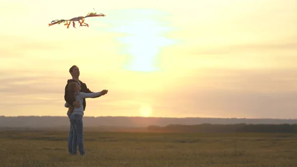 Папа с дочкой играют с воздушным змеем на закате в парке. Счастливая семья. Семейная игра на открытом воздухе. Папа и здоровая дочь запускают разноцветный бумажный самолет в небо. Малыш запускает воздушного змея — стоковое фото