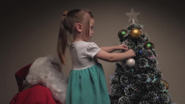 Mała dziewczynka i Święty Mikołaj powieszą piękne kulki na drzewie. Wesołych Świąt. Dziecko i Święty Mikołaj udekorować choinkę. Święta i uroczystości. Dzieci rodzinne zimowe wakacje. — Wideo stockowe