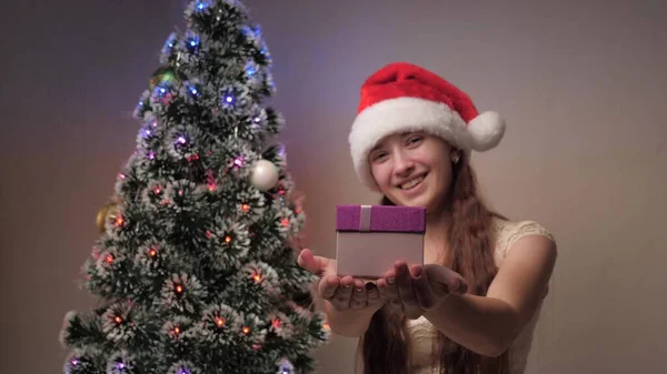 Dziewczyna w kapeluszu Świętego Mikołaja trzyma prezent obok choinki. Nastolatek trzyma pudełko prezentów przed kamerą, raduje się i uśmiecha. Rodzinne wakacje i uroczystości, zimowy odpoczynek. Wesołych Świąt. — Zdjęcie stockowe