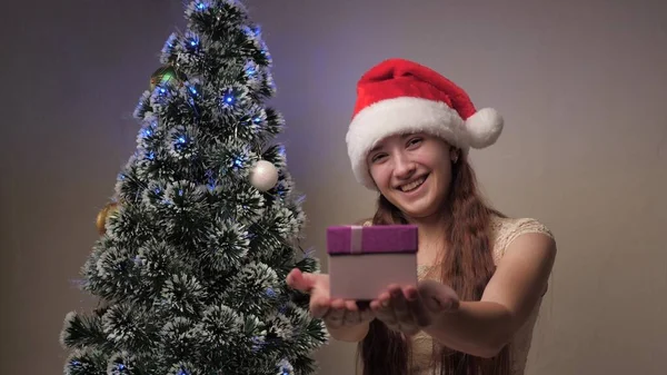 一个戴着圣诞老人帽子的女孩在圣诞树旁拿着礼物。青少年们把礼品盒分发给相机、欢乐和微笑。孩子们的节日和庆祝活动，寒假。圣诞快乐. — 图库照片