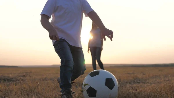 Happy family and child have fun playing a soccer ball in park. The kid kicks ball. Mom and daughter play football on field in rays of the sunset. Teamwork of the child and mom. Healthy sports family