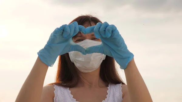 En ansiktslös kvinna i skyddsmask och handskar visar en hjärtsymbol med händerna. Ung kvinna älskar läkare. Pandemi, vid 19 års ålder. Kvinnlig läkare visar fingrar hjärta och kärlek för patienter. — Stockfoto