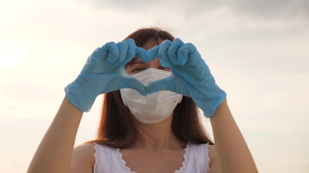 Una mujer sin rostro con una máscara protectora y guantes médicos muestra un símbolo del corazón con sus manos. La joven ama a los médicos. Pandemia, covidio 19. Doctora muestra los dedos del corazón y el amor por los pacientes. — Vídeos de Stock