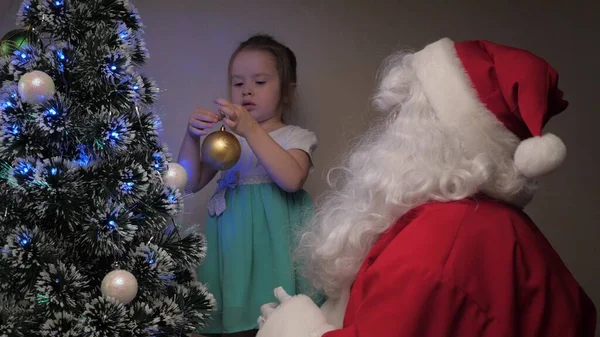 一个小女孩和圣诞老人在树上挂着漂亮的球。孩子和圣诞老人装饰着圣诞树.假期和庆祝活动。孩子们的寒假。圣诞快乐. — 图库照片