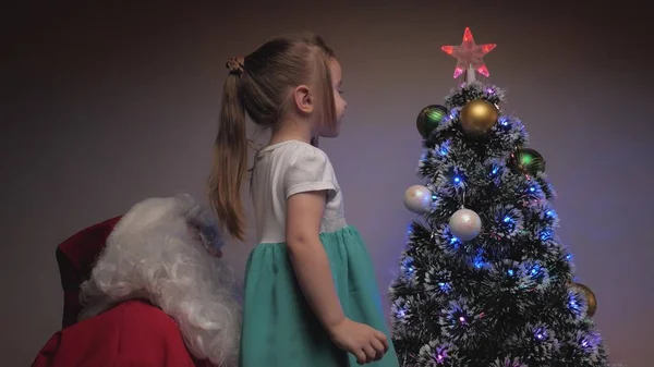 小さな女の子とサンタクロースは木の上に美しいボールを掛けます。メリークリスマス。子供とサンタクロースはクリスマスツリーを飾る。休日やお祝い。家族の子供たち冬休み. — ストック写真