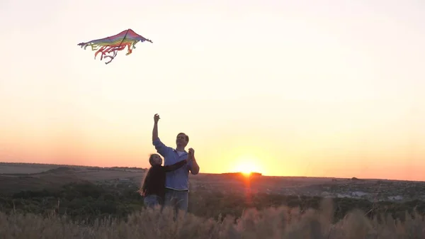 Dad plays with his daughter together and launch a kite in the park at sunset. Happy family. The child and father have fun flying kite in the air. Family launching colorful paper airplanes into sky — Stock Photo, Image