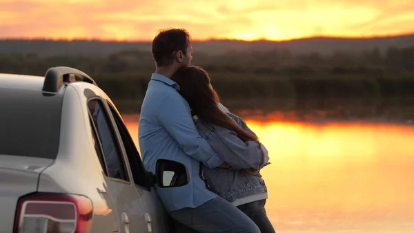 Szczęśliwi kochankowie podróżujący mężczyzna i kobieta stoją obok samochodu i podziwiają piękny zachód słońca na plaży. Turyści podróżują samochodem, przytulają się, podziwiają wschód słońca, rzekę. Wolni podróżnicy, turyści. Podróże rodzinne samochodem. — Zdjęcie stockowe