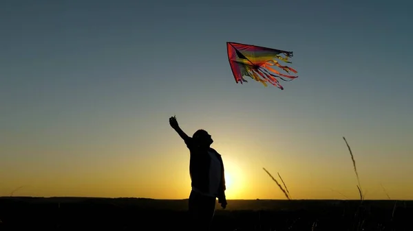 Una chica feliz corre con una cometa en sus manos a través del campo en los rayos de la puesta del sol. Un niño sano sueña con la libertad, el vuelo. El chico juega afuera en el parque. Adolescente quiere ser piloto — Foto de Stock