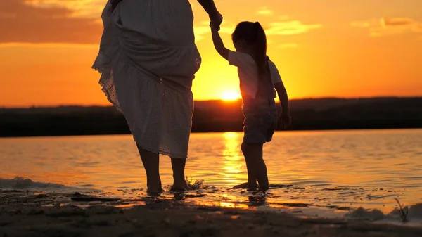 Feliz familia, mamá camina con el niño descalzo en el agua, uniendo las manos en los rayos de una hermosa puesta de sol. Madre con su hija relajándose en la playa. Silueta de una mujer y un niño en la playa al atardecer. —  Fotos de Stock