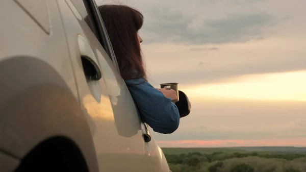 Una viaggiatrice donna tiene in mano una tazza di metallo con un caffè caldo e guarda il tramonto dal finestrino dell'auto. Autista turistico sta riposando bere il tè dalla tazza al sole. Libertà di viaggio e turismo in auto. — Foto Stock