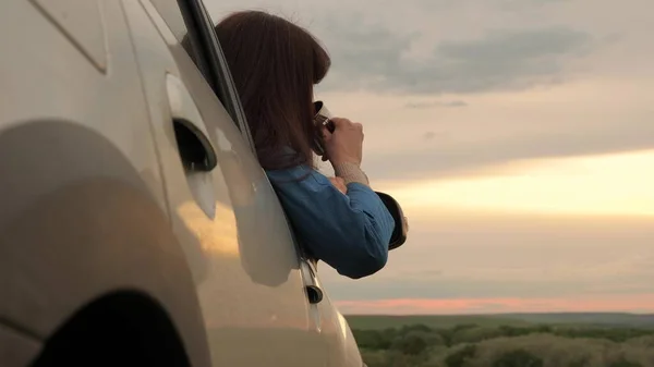 Una viaggiatrice donna tiene in mano una tazza di metallo con un caffè caldo e guarda il tramonto dal finestrino dell'auto. Autista turistico sta riposando bere il tè dalla tazza al sole. Libertà di viaggio e turismo in auto. — Foto Stock