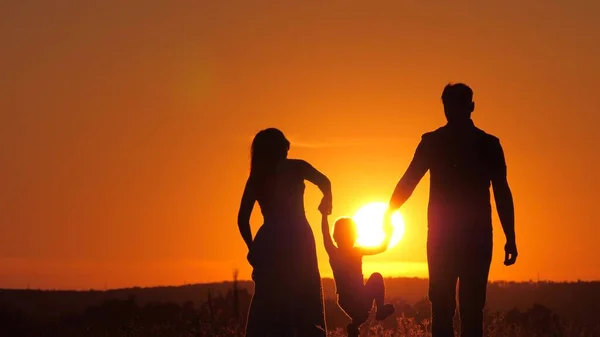 Lycklig familj, lilla dotter hoppar, håller hand med pappa och mamma i parken i solen. Barnet leker med pappa och mamma på fältet i solnedgången. Gå med ett litet barn i naturen. barndom — Stockfoto