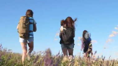 Sırt çantalı bir grup gezgin, anne ve kızları, küçük bir çocuk, yazın çiçeklerle dolu bir çayırda seyahat ediyorlar. Turistlerin takım çalışması. Yürüyüş. Mutlu bir aile. Kadın turist takımı..