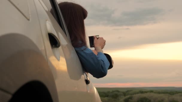 Μια γυναίκα ταξιδιώτης κατέχει μια μεταλλική κούπα με ζεστό καφέ στα χέρια της και κοιτάζει το ηλιοβασίλεμα από το παράθυρο του αυτοκινήτου. Τουριστικός οδηγός αναπαύεται πίνοντας τσάι από κούπα στον ήλιο. Ελεύθερη κυκλοφορία και τουρισμός με αυτοκίνητο. — Αρχείο Βίντεο