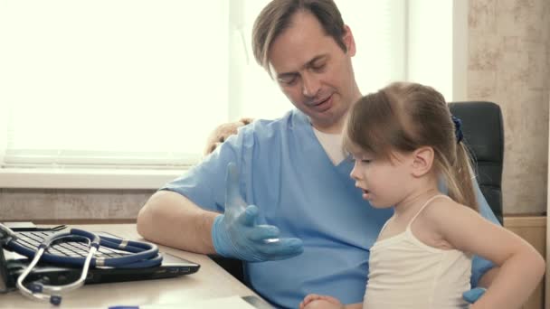 患者の赤ん坊は小児科医の膝の上に座る。かわいい女の子の子供と彼のオフィスで遊んでいる医師小児科医とラップトップで動作します。小児の治療に関する小児検査の概念. — ストック動画
