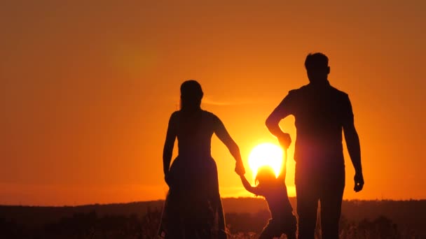 Mutlu aile, küçük kız atlıyor, parkta anne ve babasının elini tutuyor. Çocuk gün batımında anne ve babasıyla sahada oynuyor. Doğada küçük bir çocukla yürümek. çocukluk — Stok video