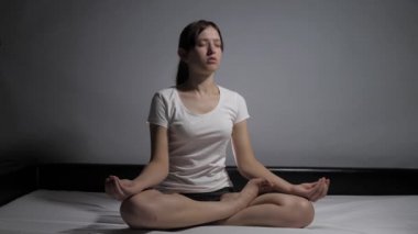 Pijamalı rahat genç bir kadın, evde gözleri kapalı yatağında oturmuş elleriyle meditasyonun tadını çıkarırken lotus pozisyonunda meditasyon yapıyor. Kız sabahları jimnastik yapıyor. Sağlık ve sükunet kavramı