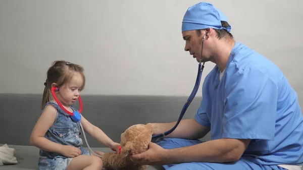 Opiekuńczy profesjonalny pediatra bawić się z małym dzieckiem w pokoju. Mała dziewczynka bawi się pluszowym miśkiem Teddy słucha go stetoskopem. Dziecko, maluch i lekarz pediatra bawią się razem — Zdjęcie stockowe