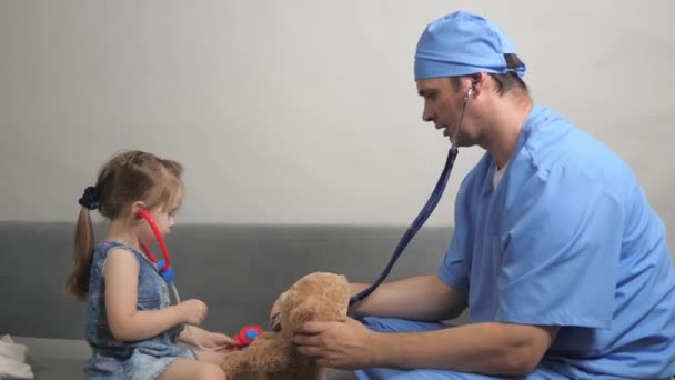 Cuidar profissional pediatra do sexo masculino brincando com a criança pequena no quarto. A menina brinca com um ursinho Teddy ouve-o com estetoscópio. Criança, criança e pediatra brincam juntos — Vídeo de Stock