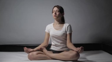 Pijamalı rahat genç bir kadın, evde gözleri kapalı yatağında oturmuş elleriyle meditasyonun tadını çıkarırken lotus pozisyonunda meditasyon yapıyor. Kız sabahları jimnastik yapıyor. Sağlık ve sükunet kavramı