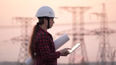 Beyaz kasklı bir kadın güç mühendisi tabletteki elektrik sensörlerinden elde edilen verileri kullanarak güç hattını kontrol ediyor. Gün batımında yüksek voltajlı elektrik hatları. Dağıtım ve elektrik tedariği. temiz enerji