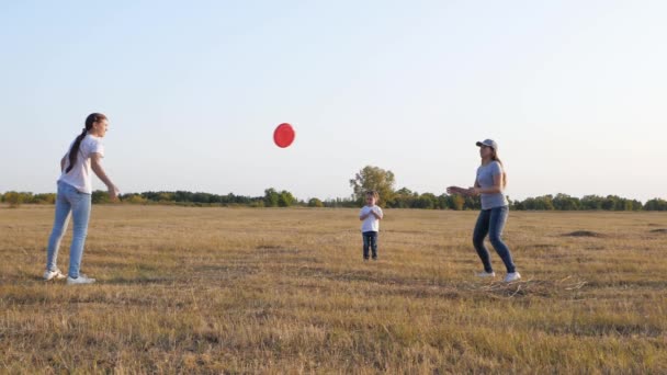 Heureux famille, mère et filles jouent, jetant un disque rouge volant les uns aux autres dans le parc. Pique-nique familial avec enfants. Les filles jettent une soucoupe volante les unes aux autres dans le parc. — Video