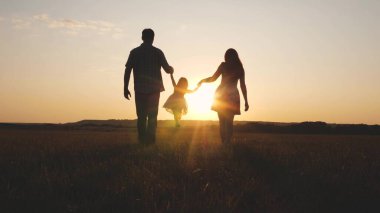 Çocuk gün batımında anne ve babasıyla sahada oynuyor. Aile ve çocukluk. Küçük kız, güneşin arka planında anne ve babasının elini tutarak zıplıyor. Doğada küçük bir çocukla yürümek..