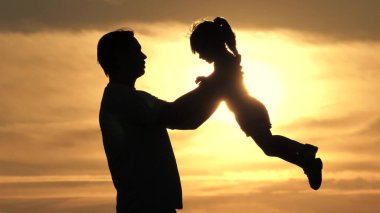 Bir baba ve çocuğun silueti, gün batımında birlikte oynuyorlar. Mutlu bir aile. Babam sevgili sağlıklı kızını kollarının arasına alıyor. Mutlu baba ve küçük çocuk parkta güneş ışığında oynuyorlar..