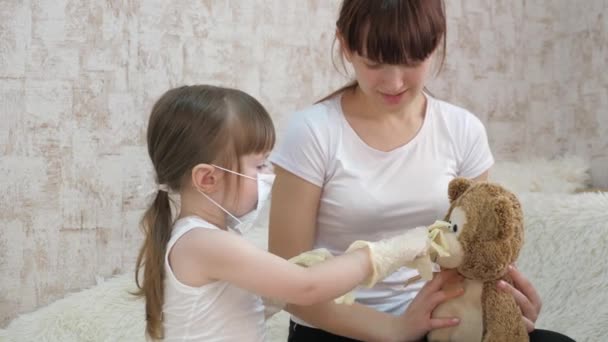 A child in a medical mask puts rubber gloves on handles and plays with his sister. Children treat a toy bear. The kid plays in hospital. Dream of becoming a doctor, nurse, veterinarian and heals sick. — Stock Video