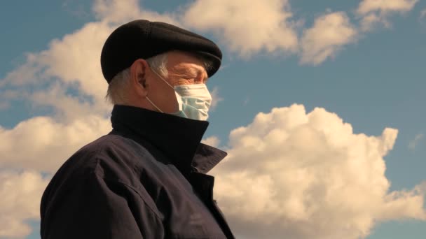 Egy férfi védőmaszkot visel az utcán. Egy idős férfi az utcán orvosi maszkban az ég hátterében. Fertőzés elleni védelem. Coronavirus-járvány. Egészségügyi ellátás, COVID-19 karantén, vírus — Stock videók