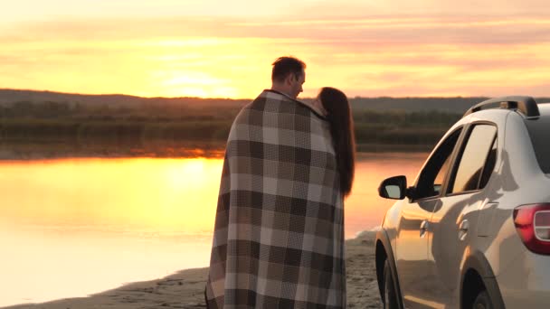 Felice viaggiatori innamorati uomo e donna si coprono con una coperta accanto alla macchina e ammirare il bellissimo tramonto sulla spiaggia. I turisti si abbracciano accanto all'auto, ammirano l'alba del fiume. Viaggiatori liberi in auto. — Video Stock