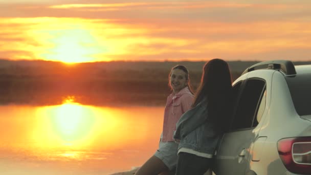 Las madres felices y la hija abrazan, viajan, se paran al lado del coche y admiran la hermosa puesta de sol en la playa. Mujeres turistas libres en coche, admirando el amanecer, río. Viajeros familiares, turistas. Viajes en familia en coche. — Vídeo de stock