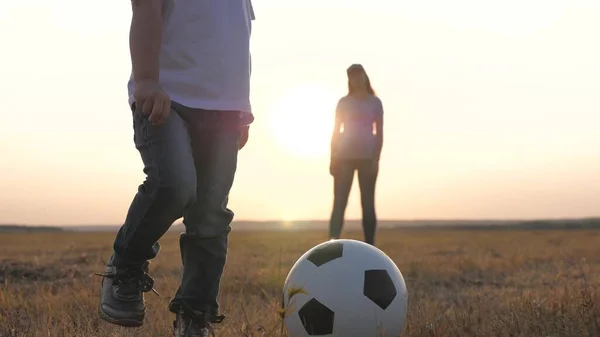 Happy family and child have fun playing a soccer ball in park. The kid kicks ball. Mom and daughter play football on field in rays of the sunset. Teamwork of the child and mom. Healthy sports family