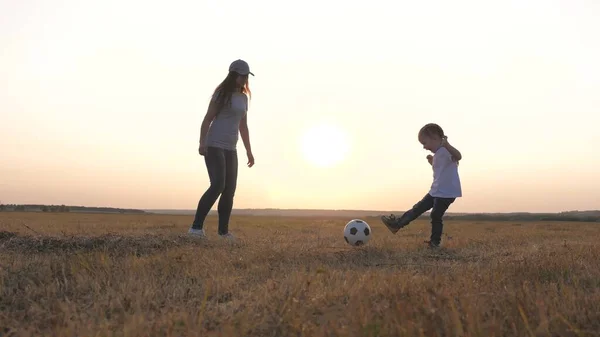 Mamma e figlia giocano a calcio sul campo a raggi del tramonto. Lavoro di squadra del bambino e la mamma. Famiglia felice e bambino si divertono a giocare a calcio nel parco. Il ragazzo prende a calci la palla. Famiglia sportiva sana — Foto Stock