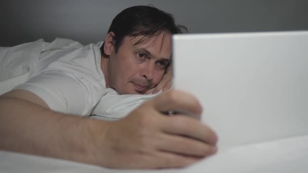 En mand hviler online hjemme om aftenen før sengetid. Tjekker post, læser nyheder i en moderne gadget. En mand derhjemme ligger på sofaen i stuen, ved hjælp af en tablet, mens shopping i en online butik. – Stock-video