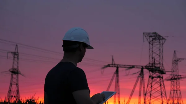 electrician works on power line. an energy engineer in a white helmet checks the power line, holds tablet in his hands. High voltage electrical lines at sunset. Distribution and supply of electricity.