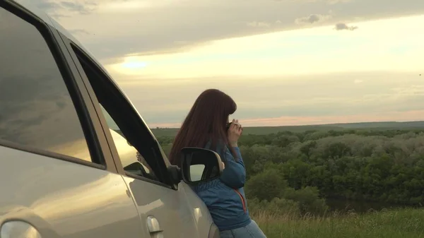 Kobieta podróżnik odpoczywa, pije poranną kawę z kubka na słońcu. Turystka trzyma kubek gorącej herbaty w rękach i patrzy na zachód słońca obok samochodu. Swoboda podróżowania i turystyki samochodem. — Zdjęcie stockowe