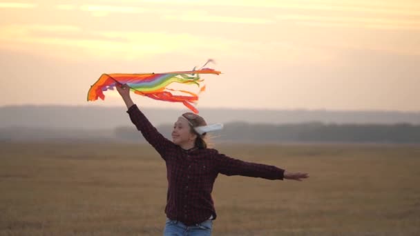 Girl wants to become a pilot. Child dreams of freedom, flight and travel. Happy child in pilots helmet runs with a multi-colored kite in his hands at sunset. Teenager playing on a plane outdoors. — Stock Video