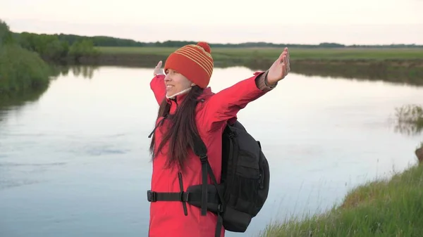 Una joven viaja caminando a la orilla del río. Mujer activa y saludable con mochila disfrutar de la naturaleza. El viajero respira aire fresco con los brazos extendidos hacia los lados y disfruta del descanso. Bioturismo, senderismo, concepto —  Fotos de Stock