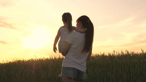 Eine Bäuerin trägt ein Kind auf dem Arm durch ein Weizenfeld. Mama spielt bei Sonnenuntergang mit ihrer kleinen Tochter. Mama spaziert mit dem Baby an der frischen Luft. Glückliche Familie von der Sonne angestrahlt — Stockfoto