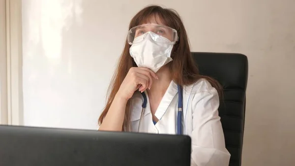 一名戴医疗面罩的女医生戴上安全眼镜，准备接诊病人。医疗概念。办公室里穿着白衣的女医生，负责笔记本电脑的研制、咨询和治疗。 — 图库照片