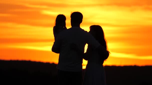 Szczęśliwa rodzina na polu podziwia zachód słońca, Dawn. Dziecko jest w ramionach ojca, żona przytula męża w promieniach ciepłego słońca. Aealthy rodzina spaceruje w parku z córką o zachodzie słońca. Mama, tata i dziecko podróżują — Wideo stockowe