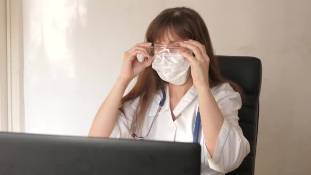 En kvinnelig lege med medisinsk maske tar på seg vernebriller og gjør seg klar til å ta imot pasienter. Medisinsk opplegg. Kvinnelig lege i hvit frakk som jobber med bærbar datamaskin, konsultasjon og behandling av – stockvideo