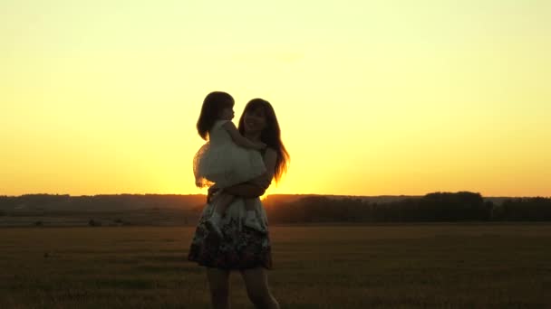 Mamãe está carregando sua amada filha saudável em seus braços, um passeio no parque no verão. A criança abraça e beija a mamã ao sol. A mãe brinca com o bebé. Uma criança com um pai anda ao pôr-do-sol. Família feliz — Vídeo de Stock